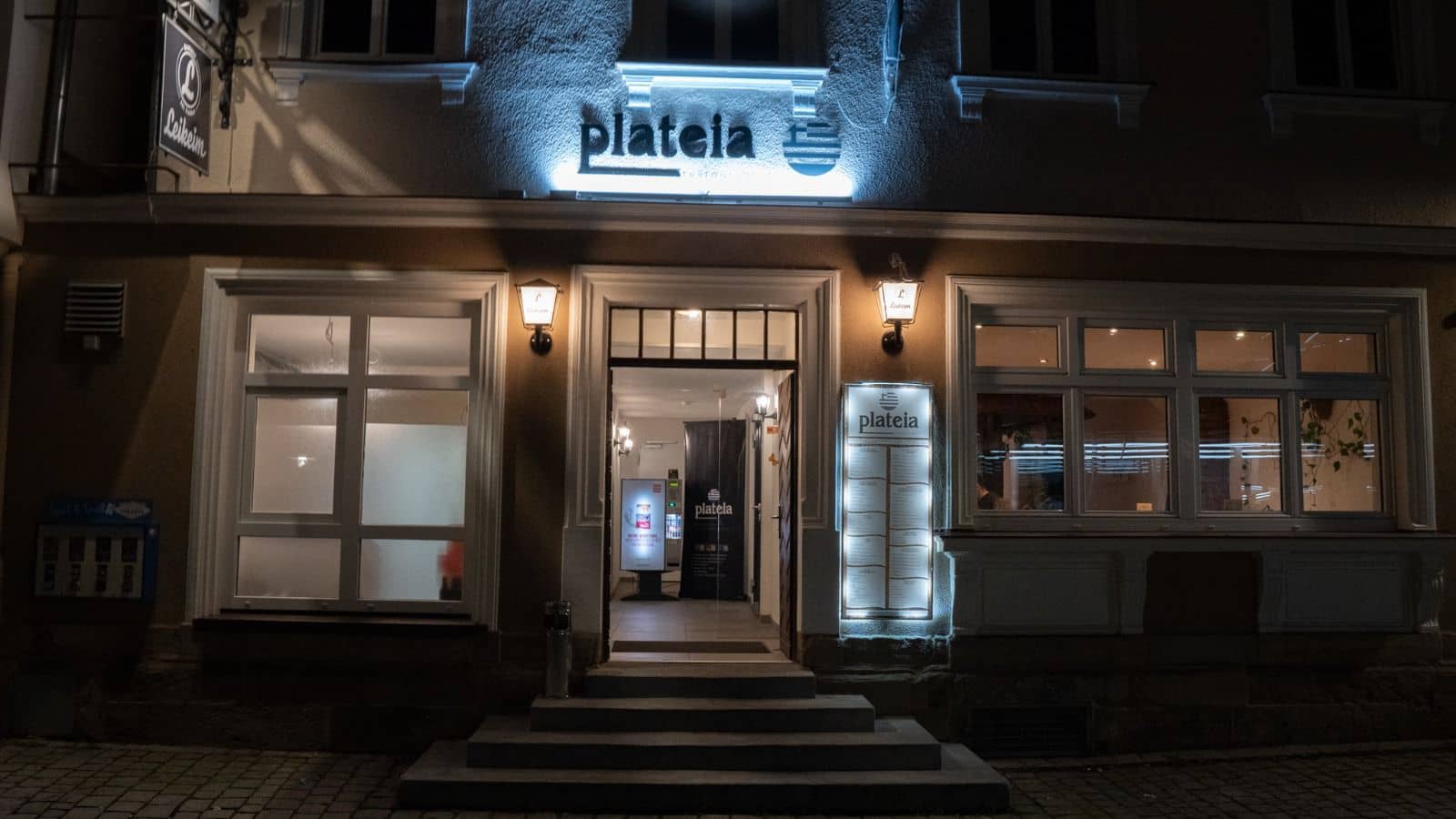 Die beleuchtete Fassade eines Restaurants namens "Plateia" bei Nacht ist zu sehen.