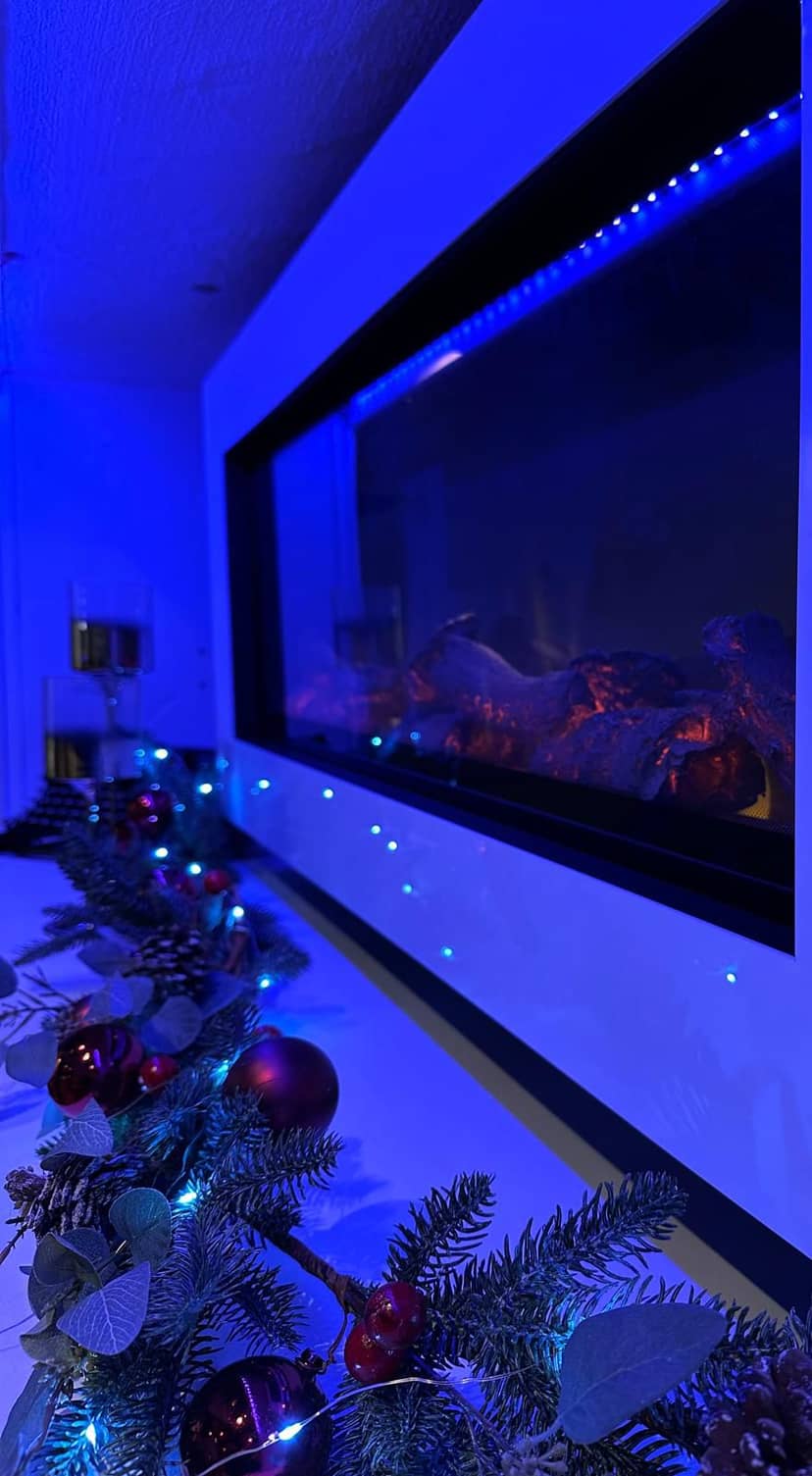 Das Foto zeigt eine weihnachtlich dekorierte Ablage mit Tannenzweigen, roten Kugeln und Lichterketten in einem Raum, der stark von blauem LED-Licht beleuchtet…